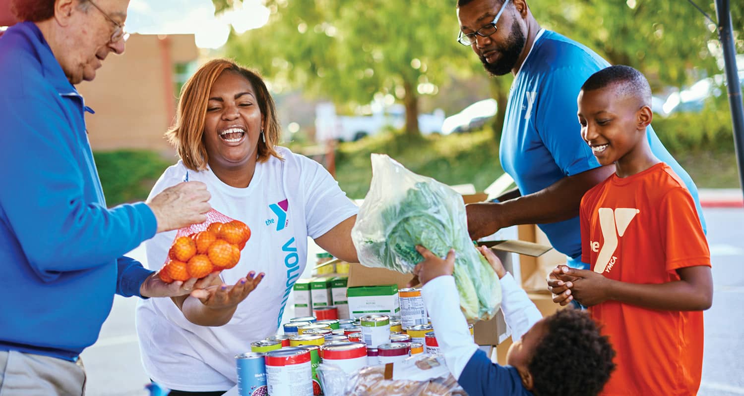 Header_community-volunteer Volunteers and children at a community food drive, smiling while distributing fresh produce and canned goods outdoors.